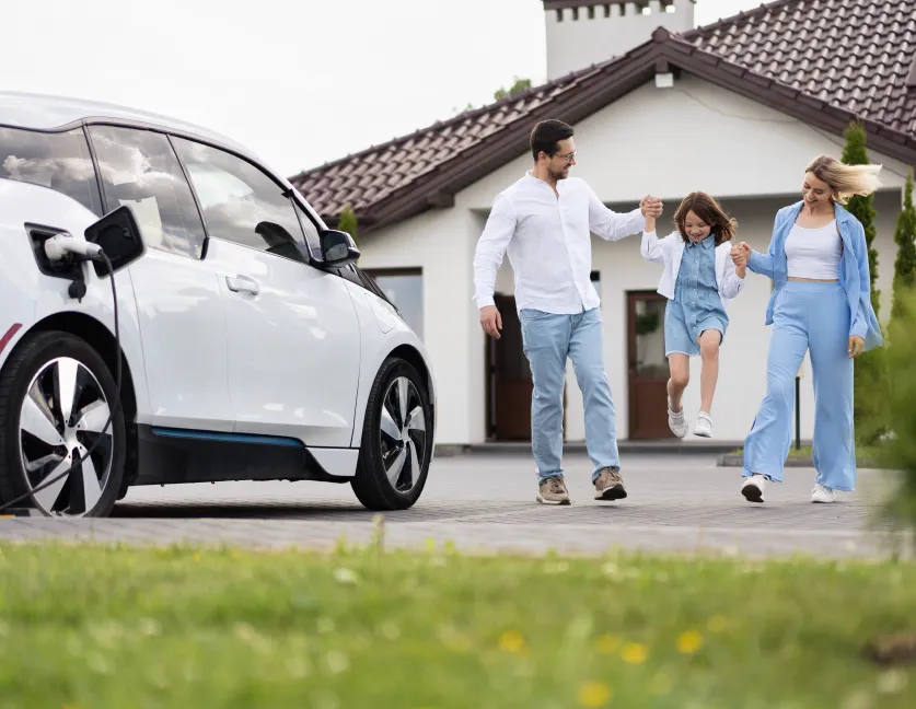 Familia feliz con un coche eléctrico cargando y su casa de fondo